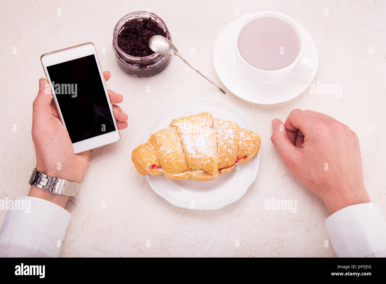 Croissant with raspberry jam, cocoa in a mug, in hand mobile phone. Business breakfast concept. Stock Photo