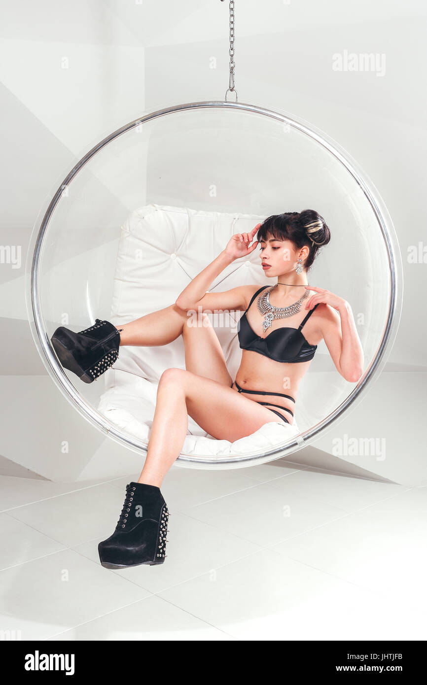Beautiful slim model woman in underwear posing siting in suspended transparent chair in white studio. Stock Photo