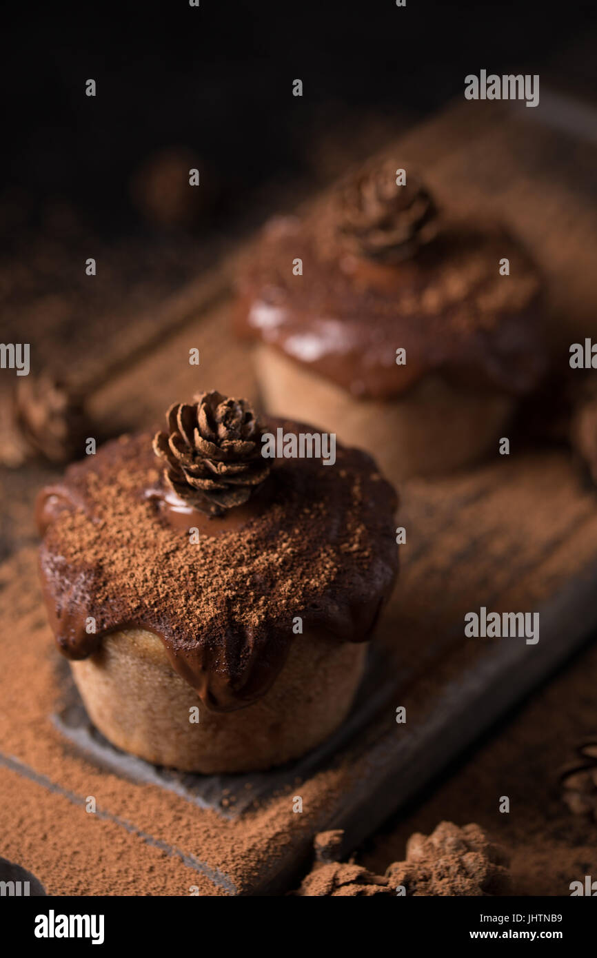 Chocolate muffins, decorated with a small cone on a dark wooden background. Low key. Cupcakes are poured with dark chocolate and cocoa powder. Cupcake Stock Photo