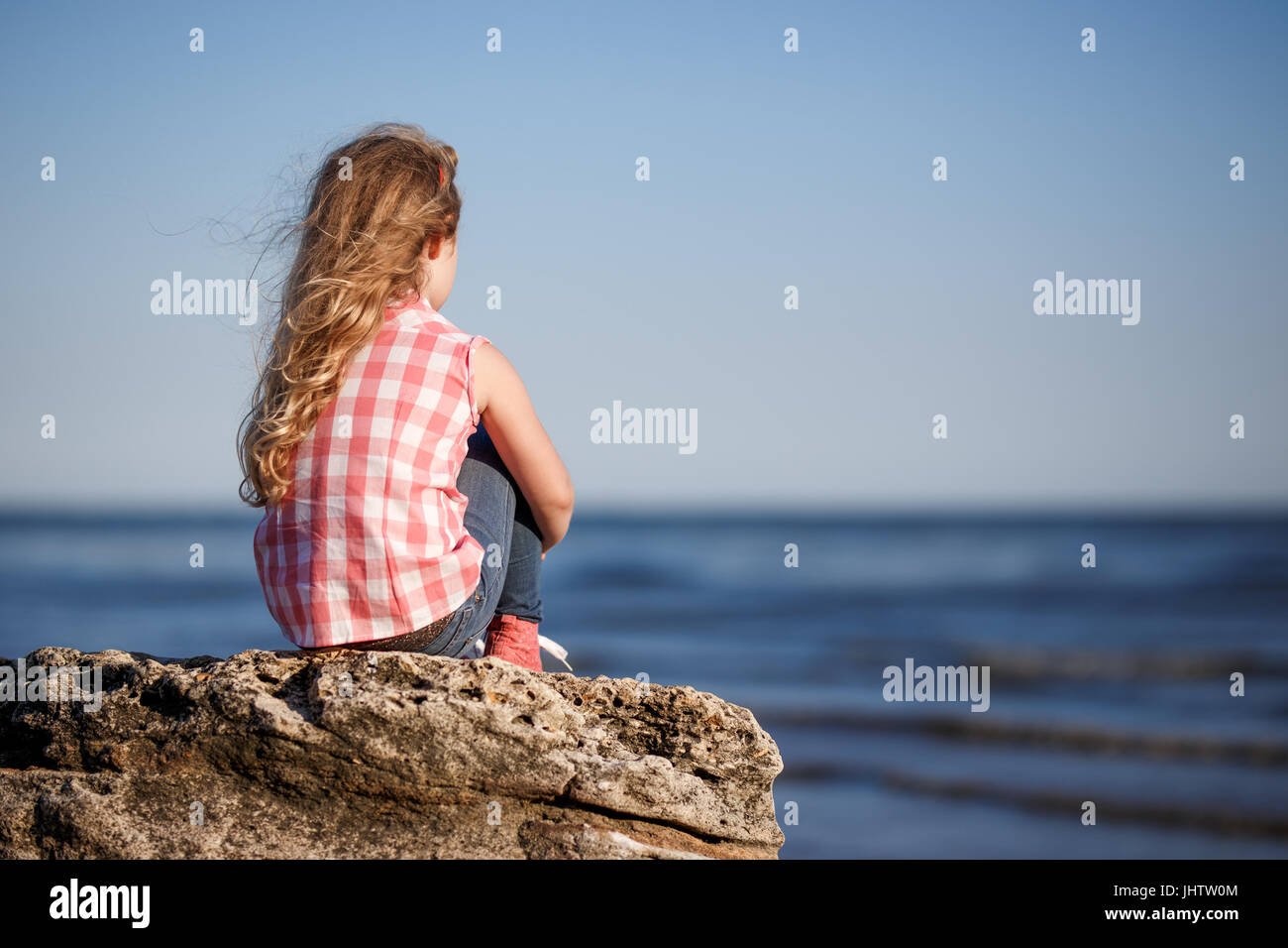 Little girl sits on a rocky shore and looks at the sea Stock Photo - Alamy