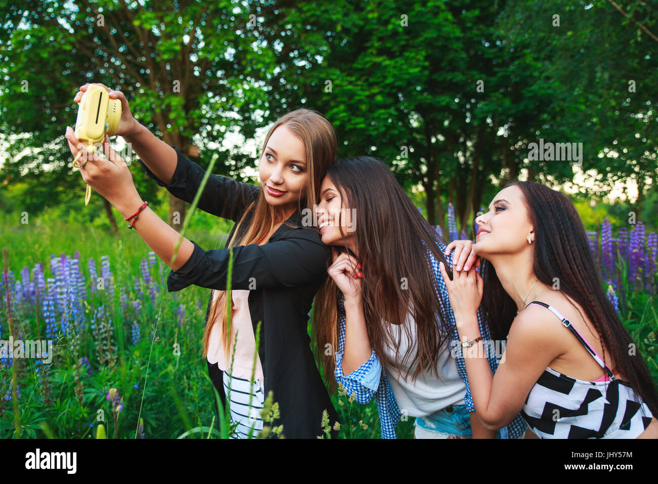 Three hipsters girls blonde and brunette taking self portrait on polaroid camera and smiling outdoor. Girls having fun together in park. Stock Photo