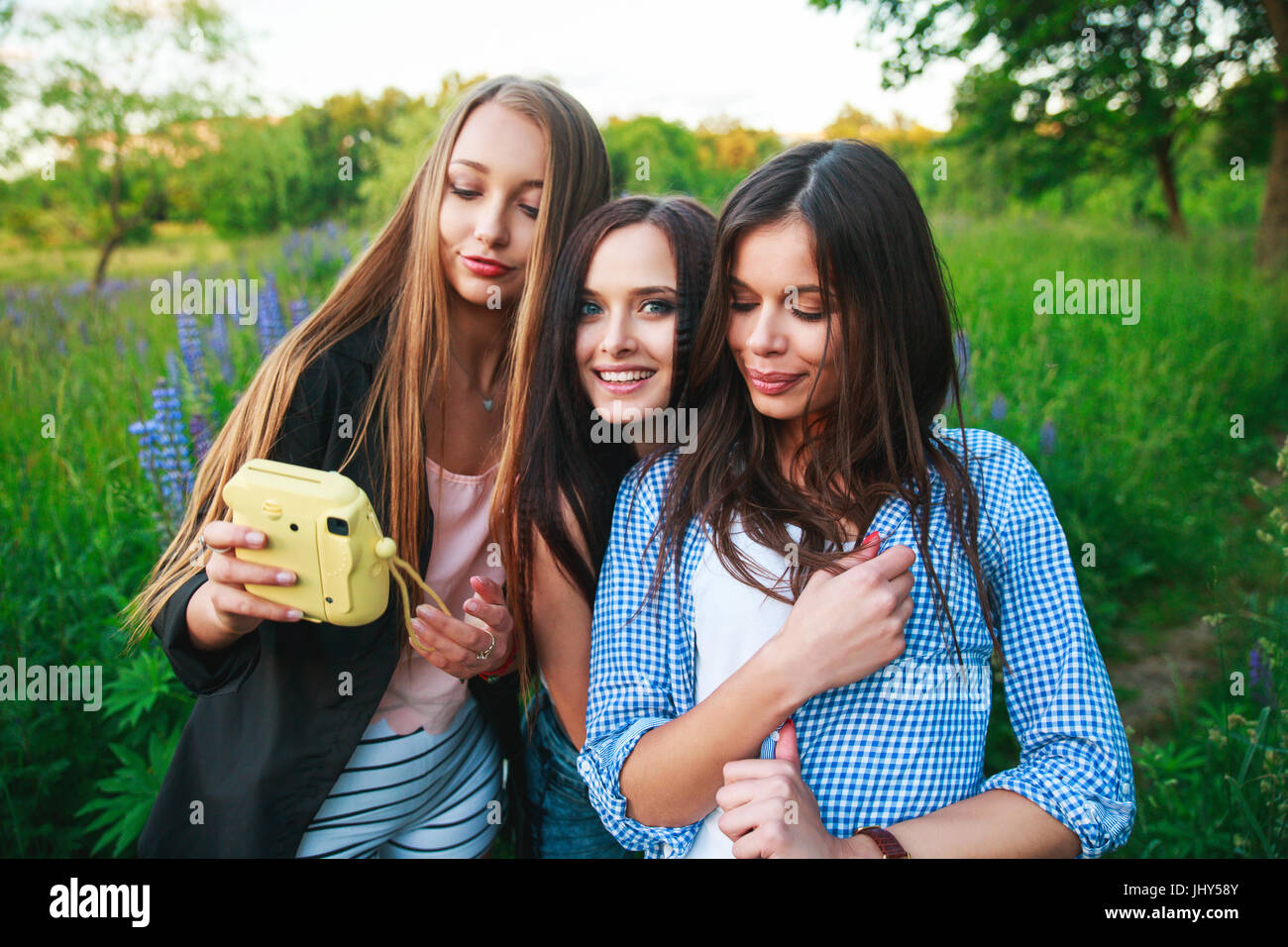 Three hipsters girls blonde and brunette taking self portrait on polaroid camera and smiling outdoor. Girls having fun together in park. Stock Photo