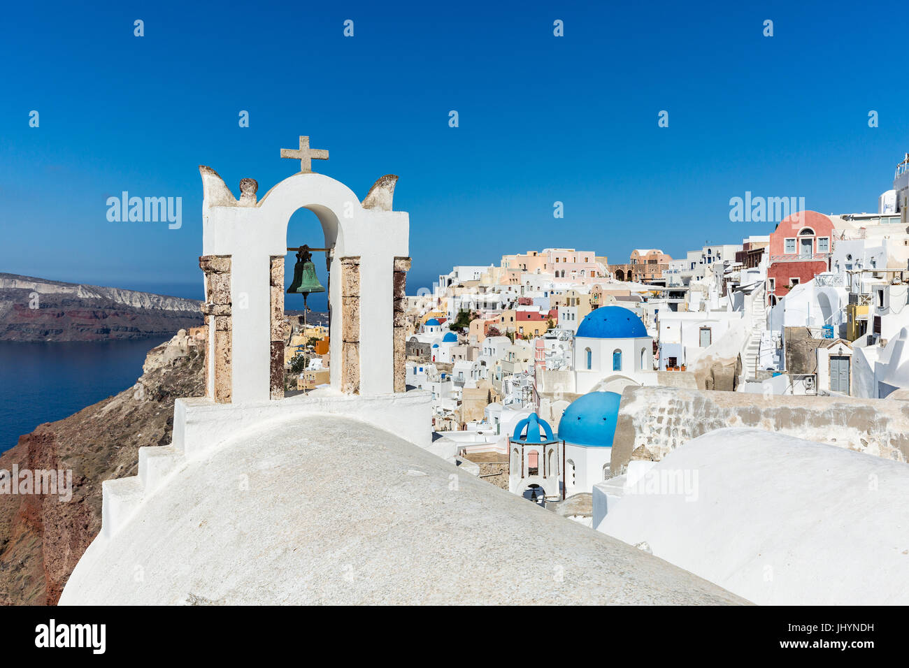 A church roof and bell with the white washed stone walls and blue church cupolas of Oia, Santorini, Greece Stock Photo
