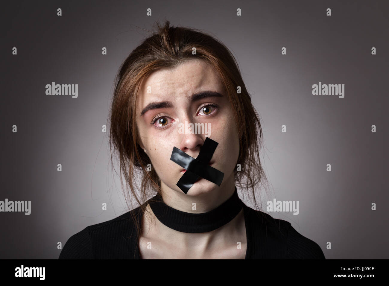 sad girl with tape on her mouth looking at camera at gray background