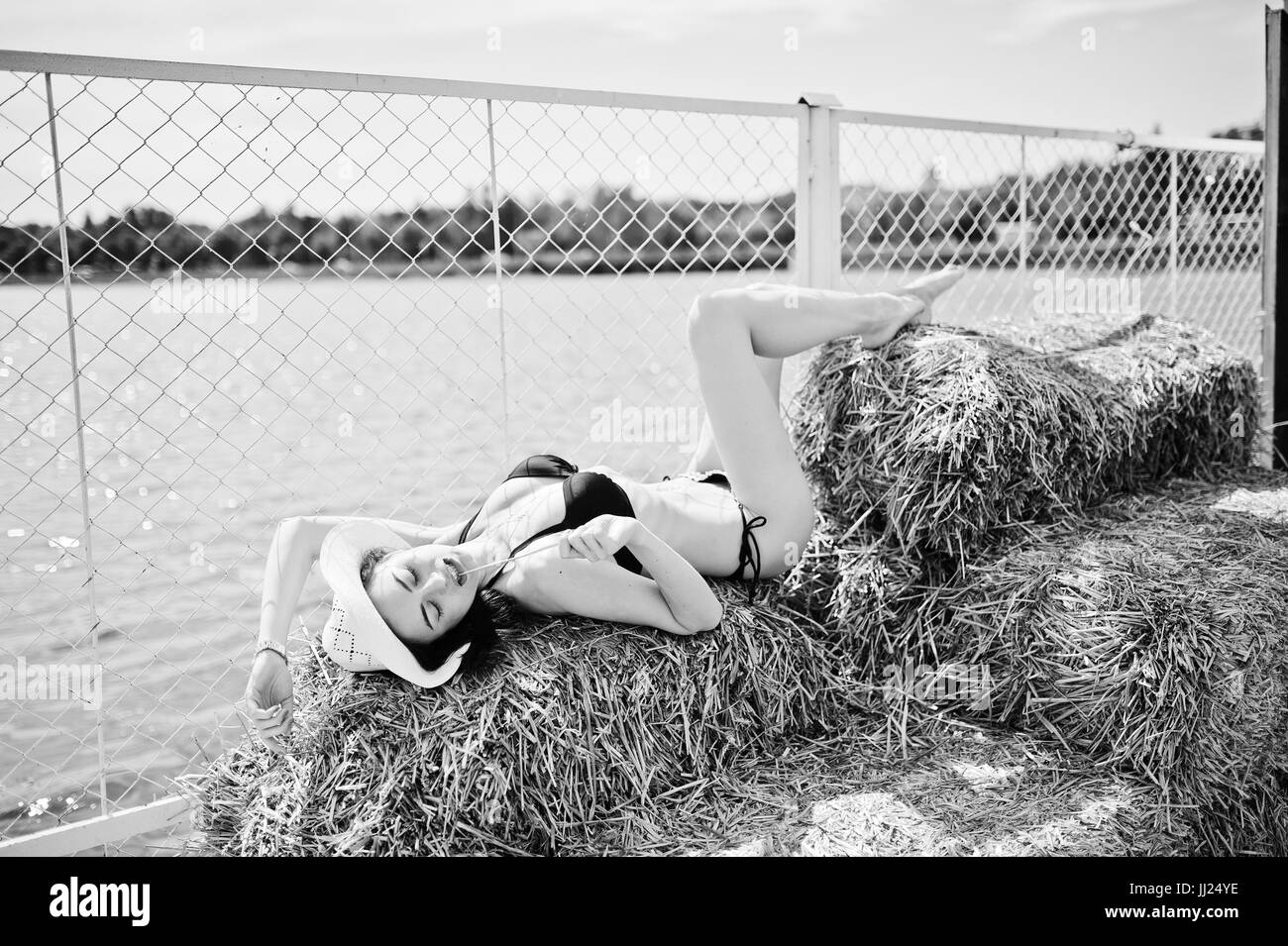 Portrait of a gorgeous girl in black bikini swimsuit posing on the hay bale with a hat by the lake. Black and white photo. Stock Photo