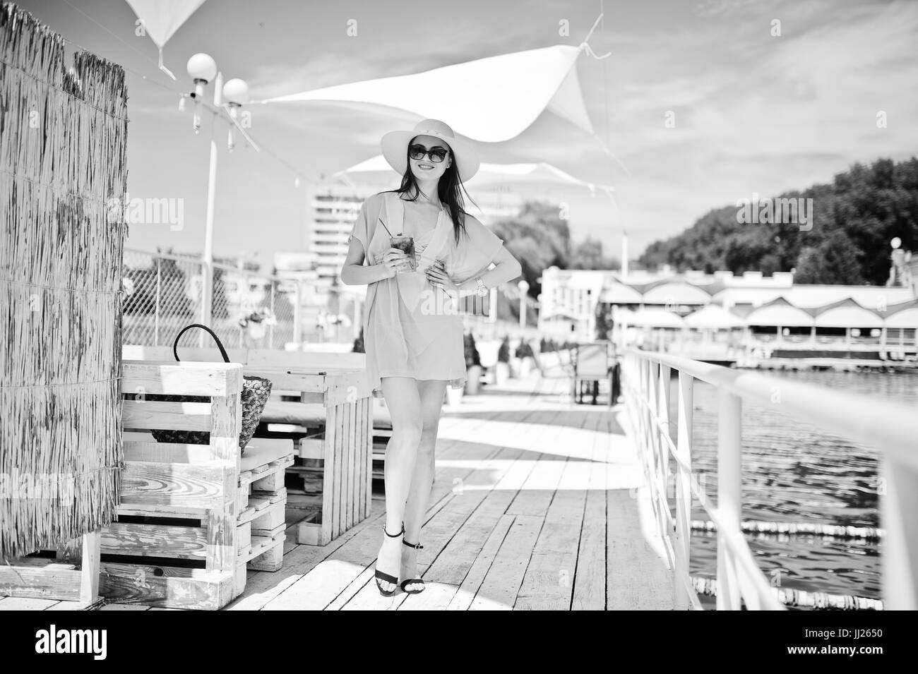 Portrait of an attractive woman in transparent turquoise dress posing with a cocktail in her hand by the lake. Black and white photo. Stock Photo