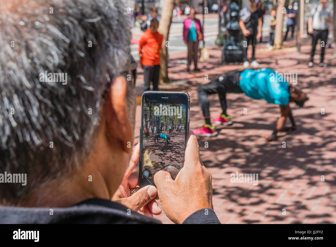 A man uses his cell phone to photograph a street performer on Market Street in San Francisco. The man is facing away and the image of the performer is Stock Photo