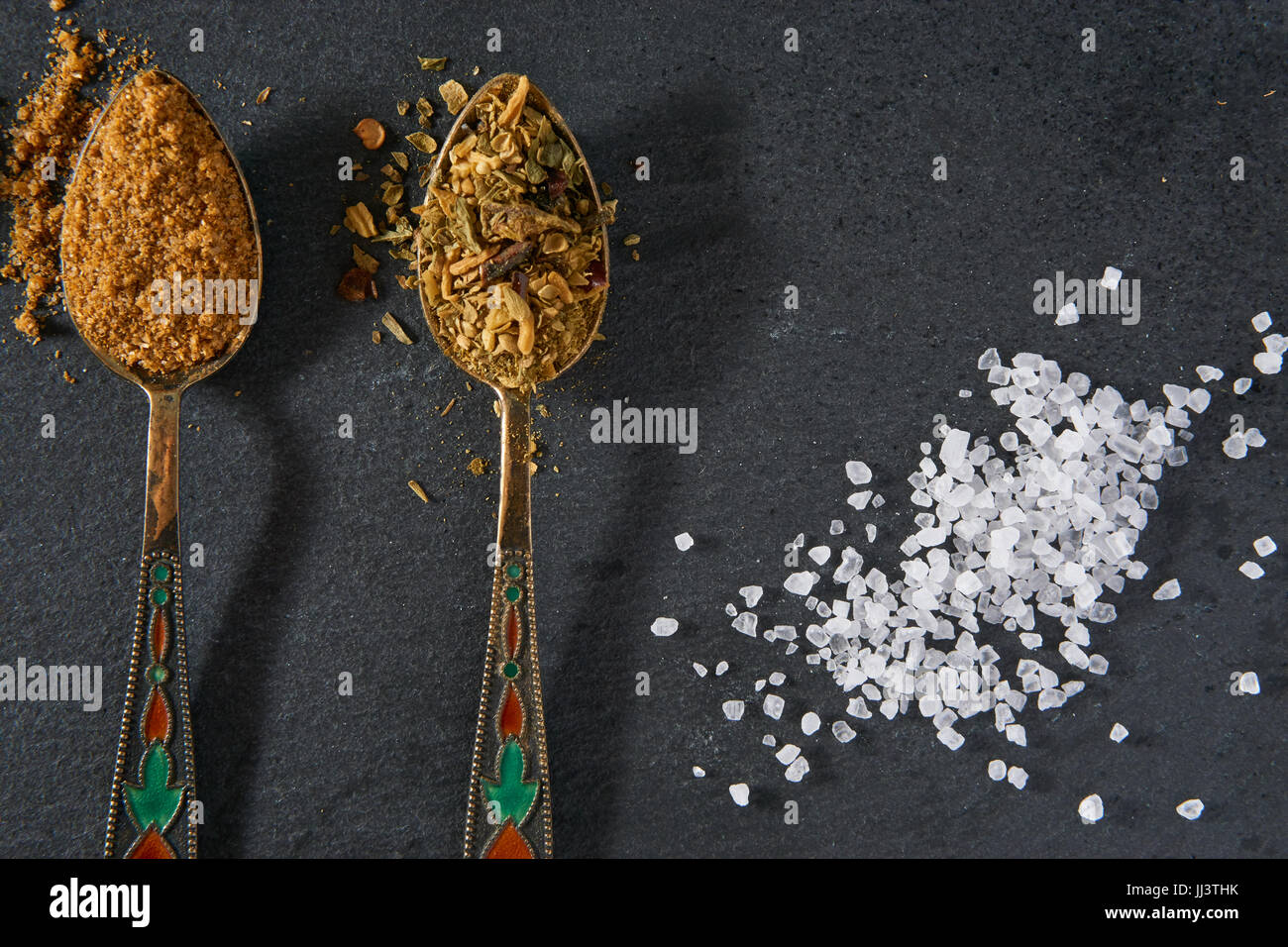 two small decorative spoons of seasoning spices and pinch of sea salt on black slate. closeup