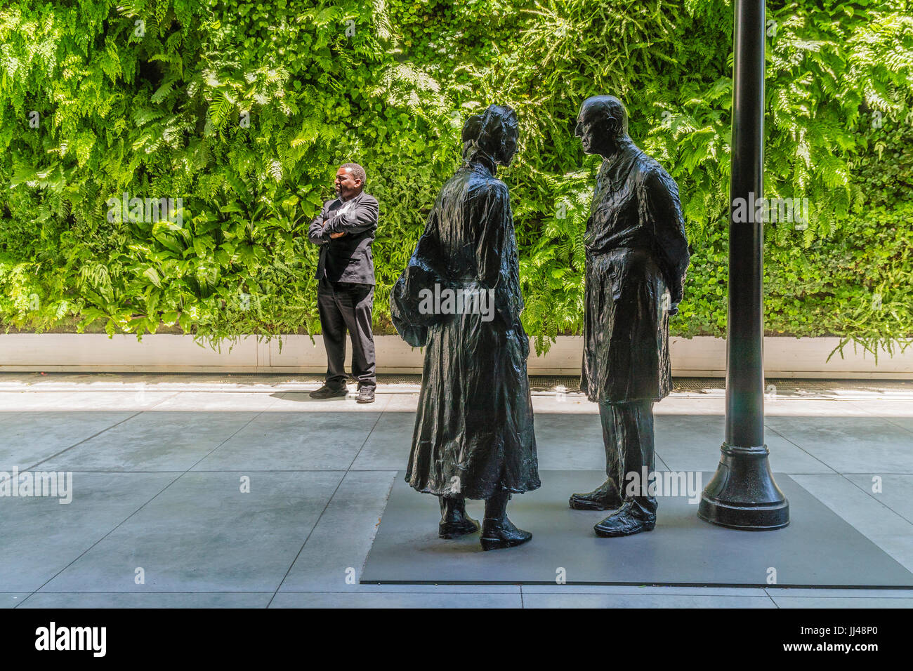 A male museum guard stands outside by a sculpture of two people with his facial profile similar to that of the male sculpture's. Stock Photo