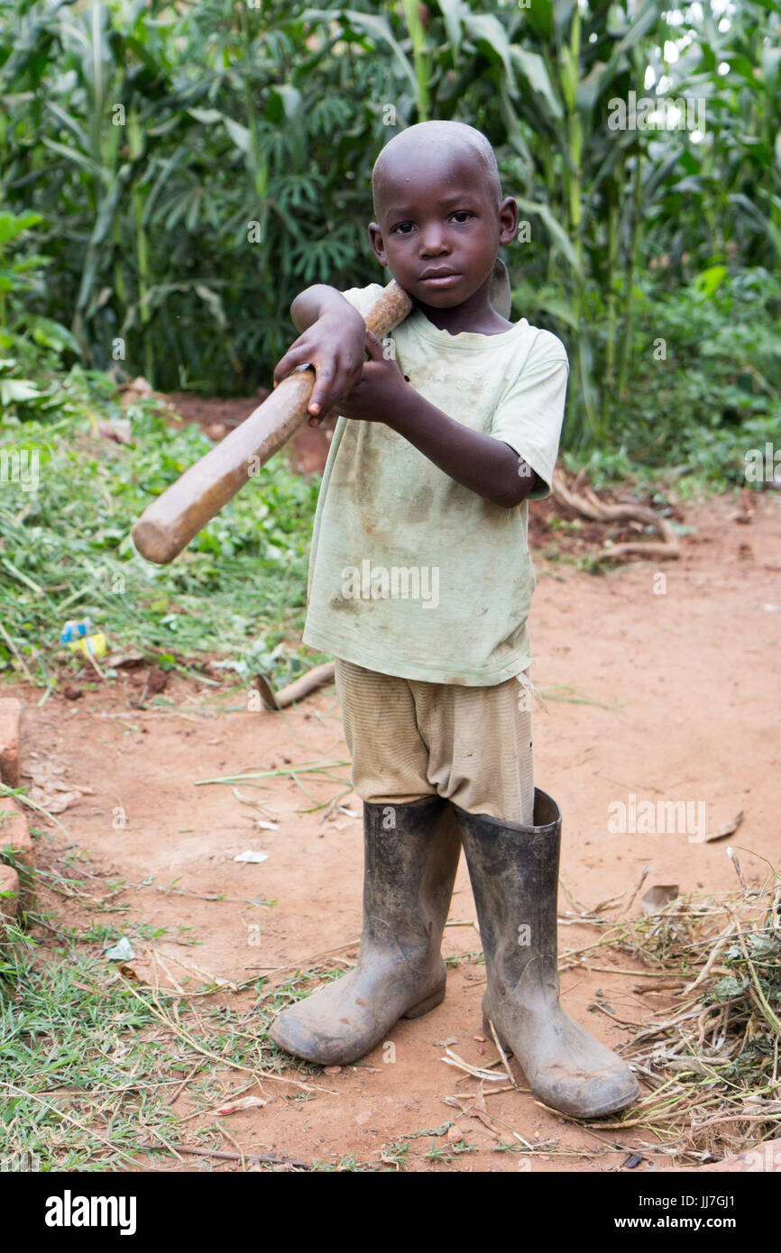 A black African Ugandan child boy holding a hoe on its shoulder. The boy is in poor rag clothes and rubber boots Stock Photo