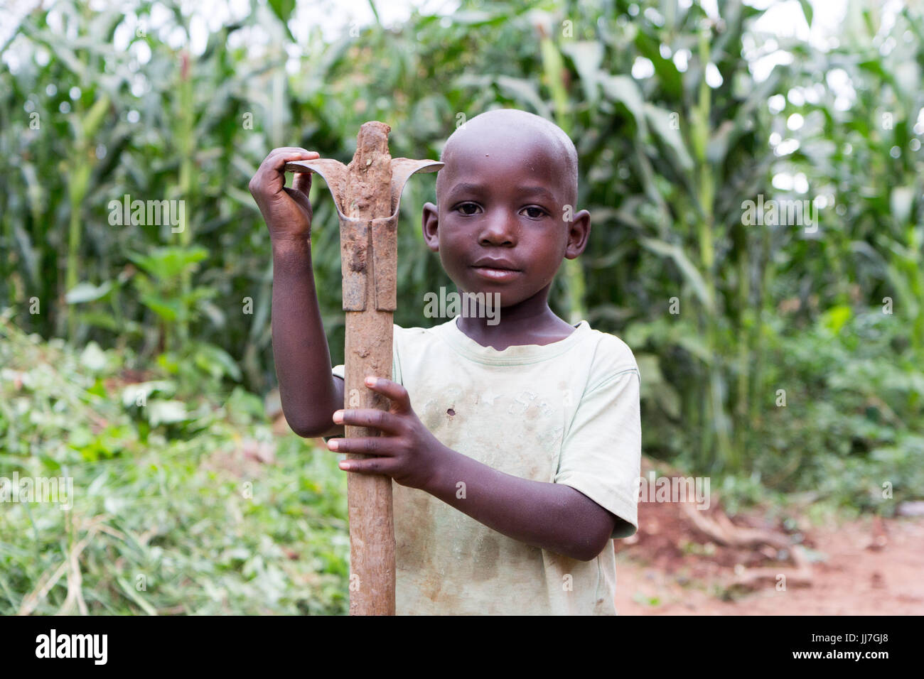 A black African Ugandan child boy holding a hoe on its shoulder. The boy is in poor rag clothes and rubber boots Stock Photo