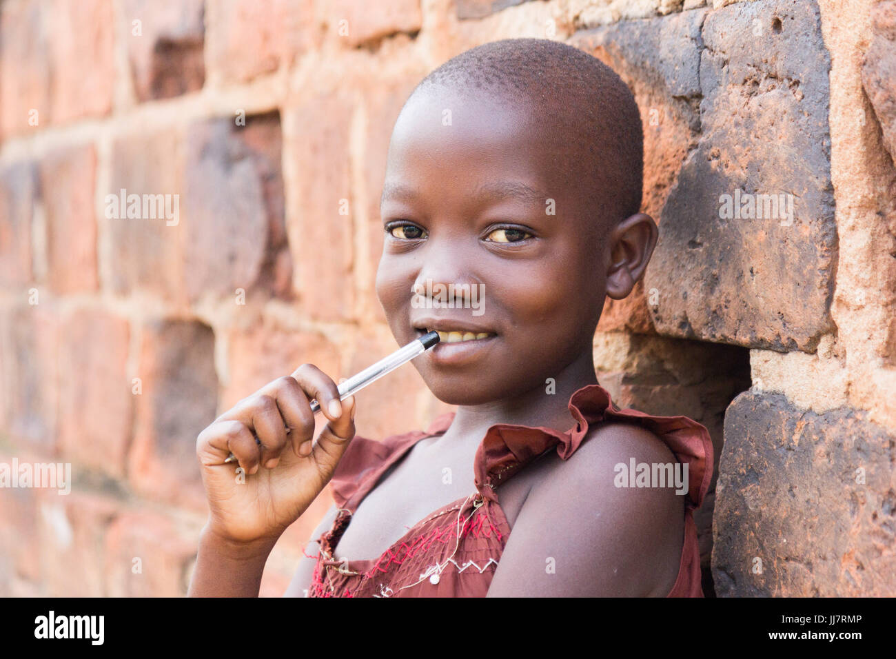 An 11-year old black Ugandan girl smiling and holding a pen against her mouth and leaning against a brick wall looking at the camera Stock Photo