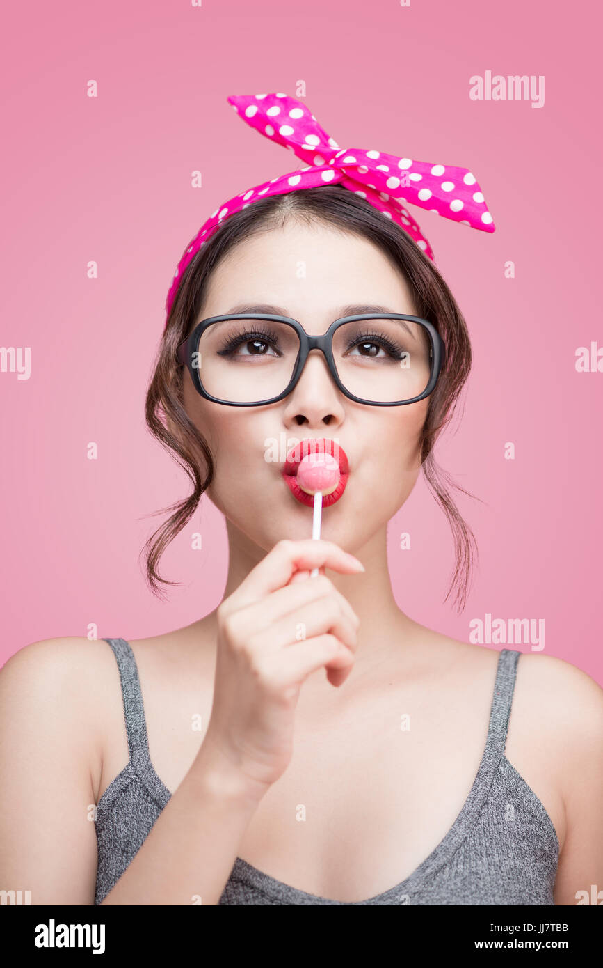 Portrait of beautiful asian woman eating heart shape lollipop, dressed and makeup in pin-up style on pink background. Stock Photo