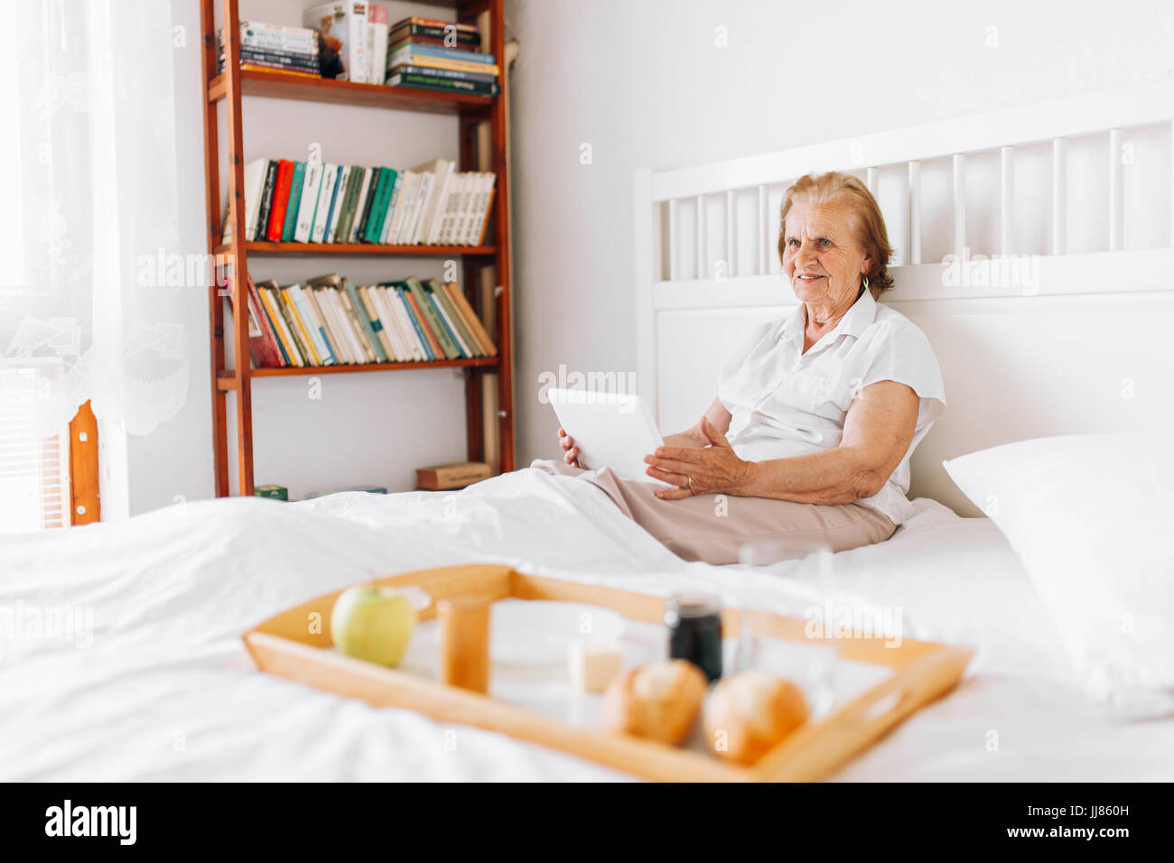 Happy elderly woman having breakfast in bed whilst using her tablet Stock Photo