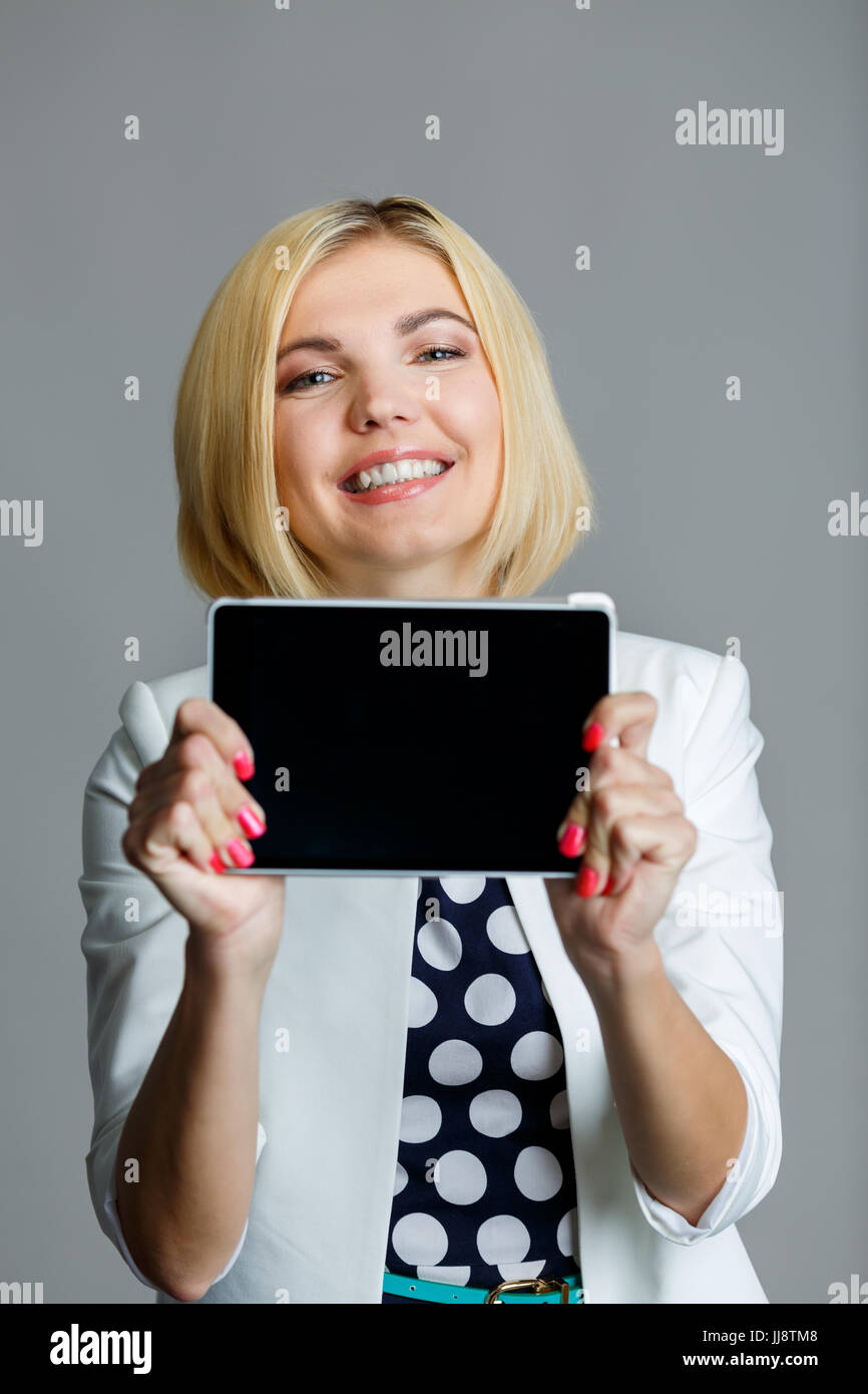 Smiling young model holds tablet with black screen on empty gray