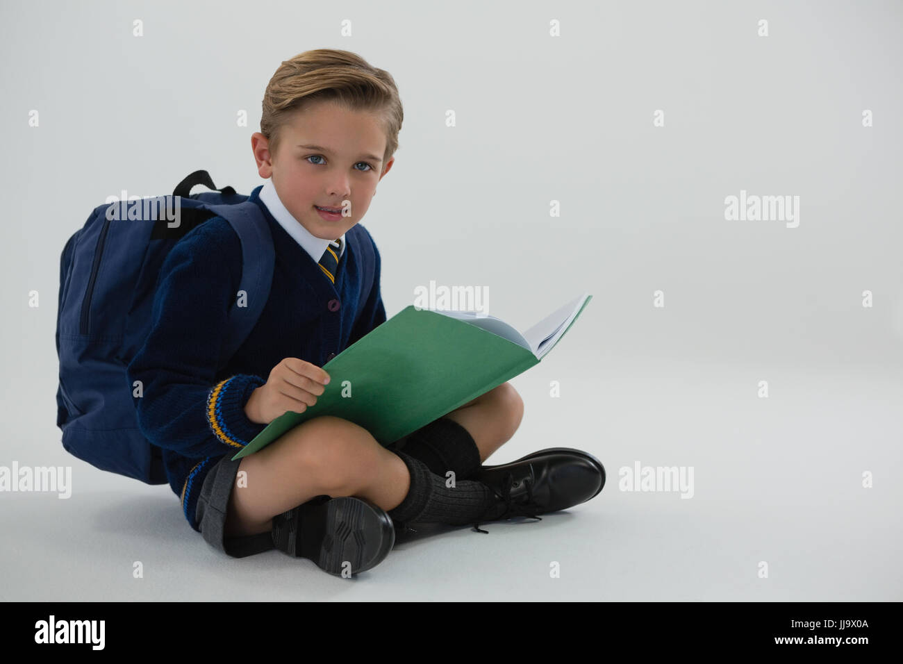 Portrait of schoolboy reading book while sitting on white background ...