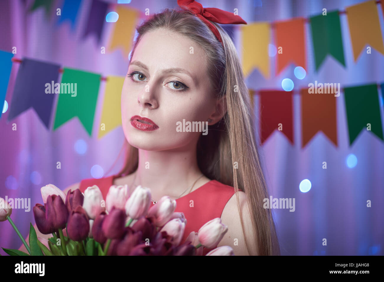 Concept: celebration, birthday. Beautiful young vintage pin up style girl standing in colorful lighted scene holding flowers. Stock Photo