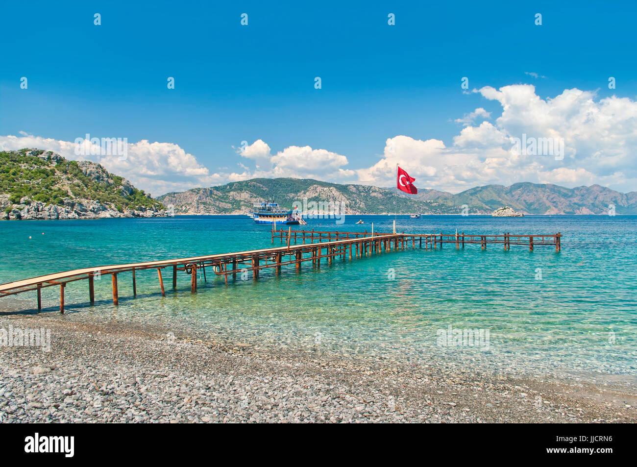 view of Aegean sea bay with tourist boats from Amos pebble beach with old rusty pier and Turkish flag on sunnt day, Marmaris, Turkey Stock Photo