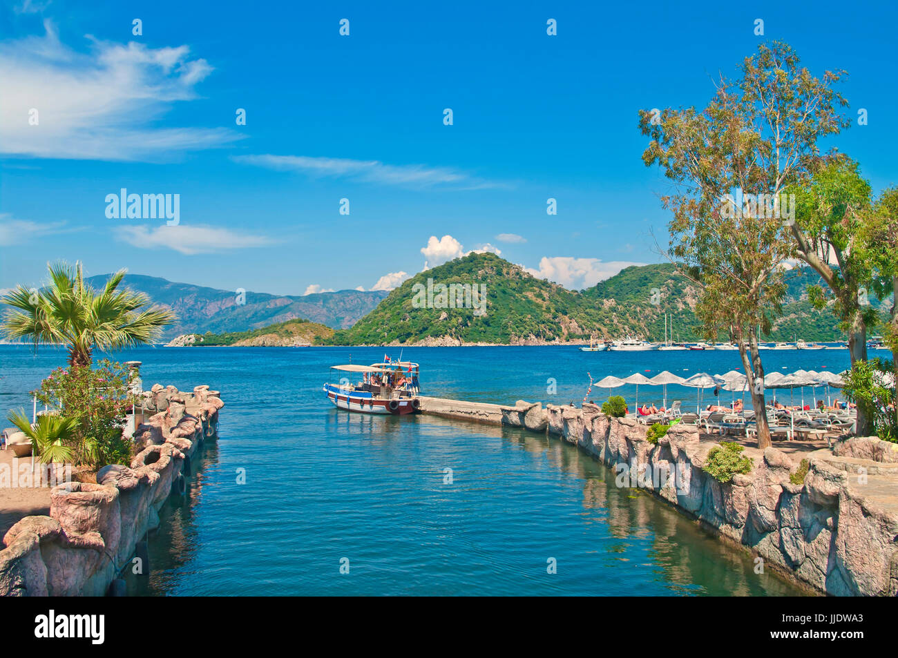 small tourist boat at Icmeler pier in Aegean sea bay with palm trees at foeground and mountains at background, Marmaris, Turkey Stock Photo