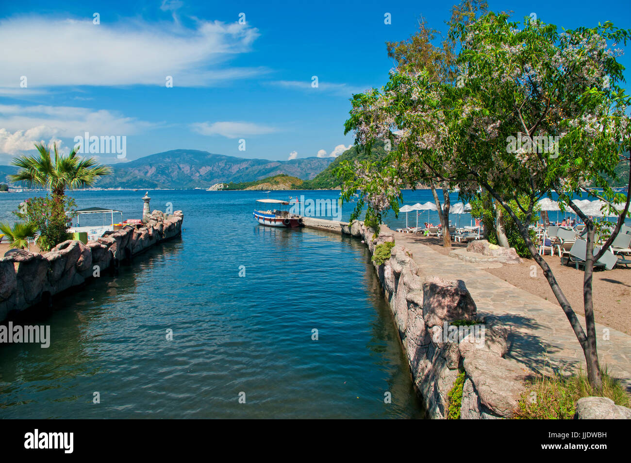 small pier with tourist boat near beach with blooming tree at foreground and mountanious islands at background, Icmeler, Marmaris, Turkey Stock Photo