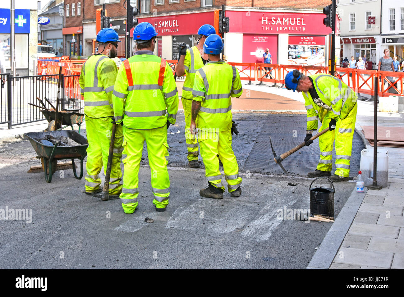 Group of workmen resurfacing town centre road one man using pick axe four others taking short break all wearing hard hats & full high visibility kit Stock Photo
