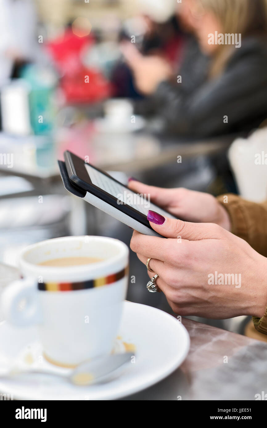 closeup of a young caucasian woman using a tablet, in the terrace of a cafe, sitting at a table, where there is a cup of coffee in the foreground Stock Photo