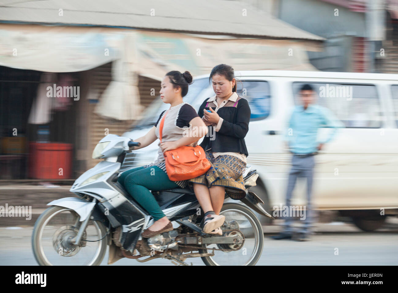 Young women ride a scooter together and check their phones on a busy street in Luang Prabang, Laos. Stock Photo