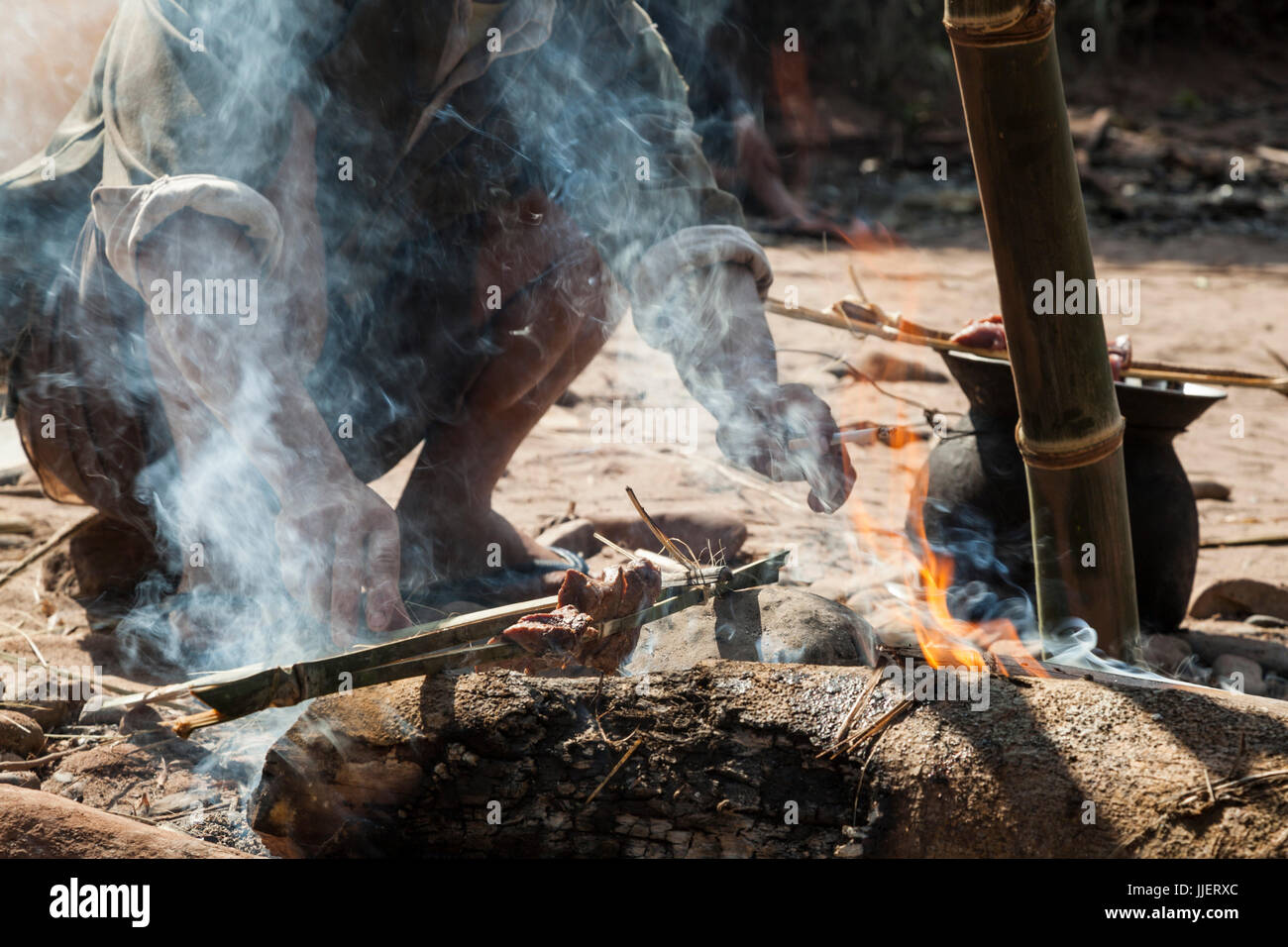 A man grills barking deer meat (Muntjacs sp.) over an open fire at his camp on the Nam Ou River in Phou Den Din National Protected Area, Laos. The area is frequented by hunters and fishermen who camp and poach wild game within park boundaries despite (or perhaps without knowledge) that this is technically illegal. Stock Photo