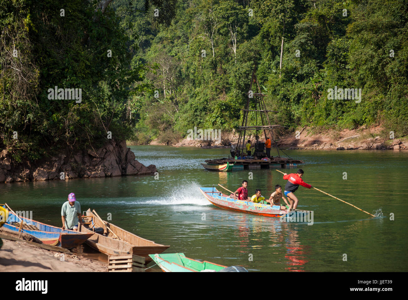 Young men speed off in a motorboat past a drill rig collecting core samples on the Nam Ou River at the planned site for Dam #7 within Phou Den Din National Protected Area, Laos. Stock Photo