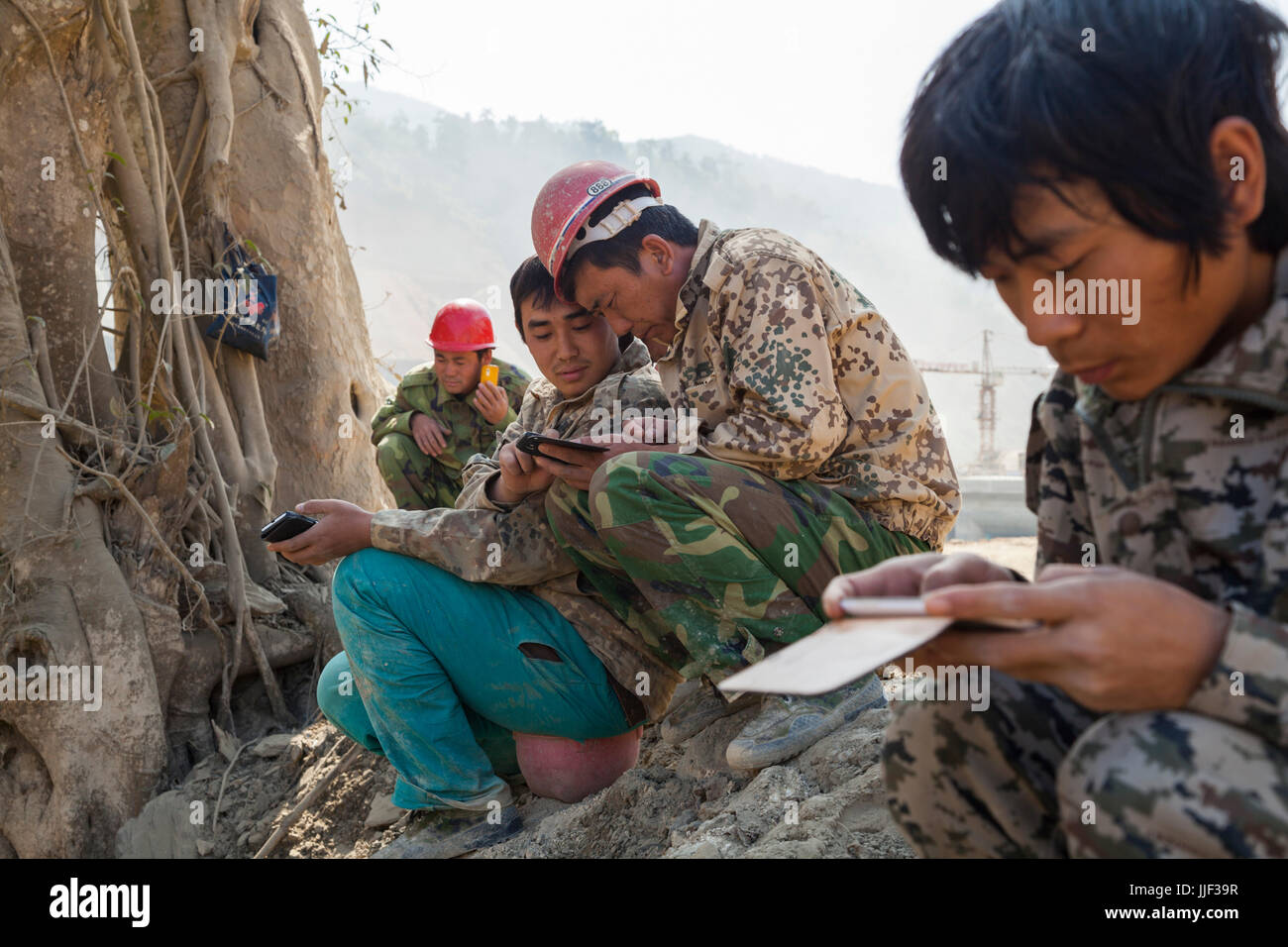 Chinese workers place calls and play video games on their smartphones at a safe distance from a planned dynamic blast at the construction site for Dam #5 on the Nam Ou River, Laos. Stock Photo