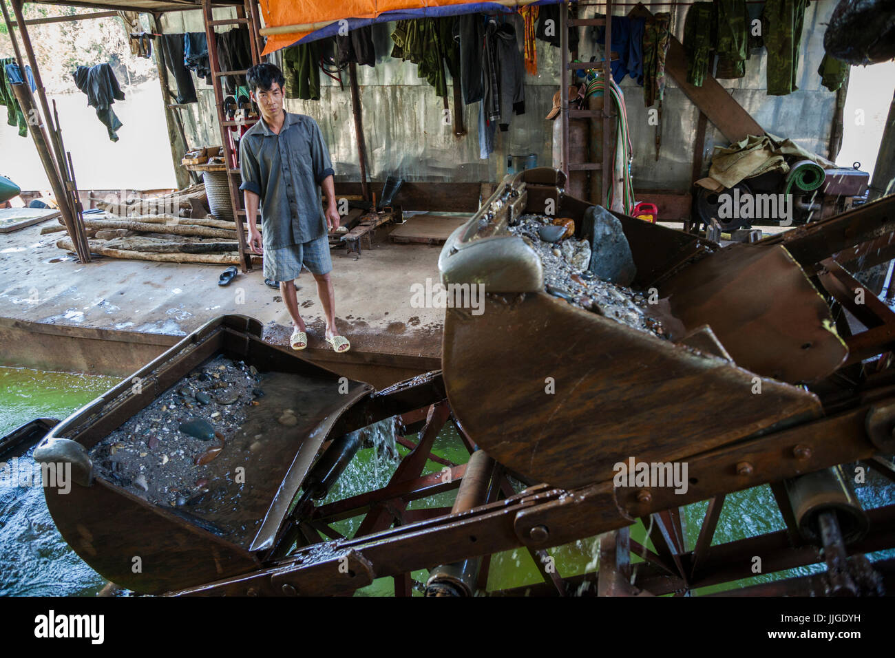 A Vietnamese man stands besides the steel buckets on an operating mechanical gold dredge floating on the Nam Ou River, Laos. Stock Photo