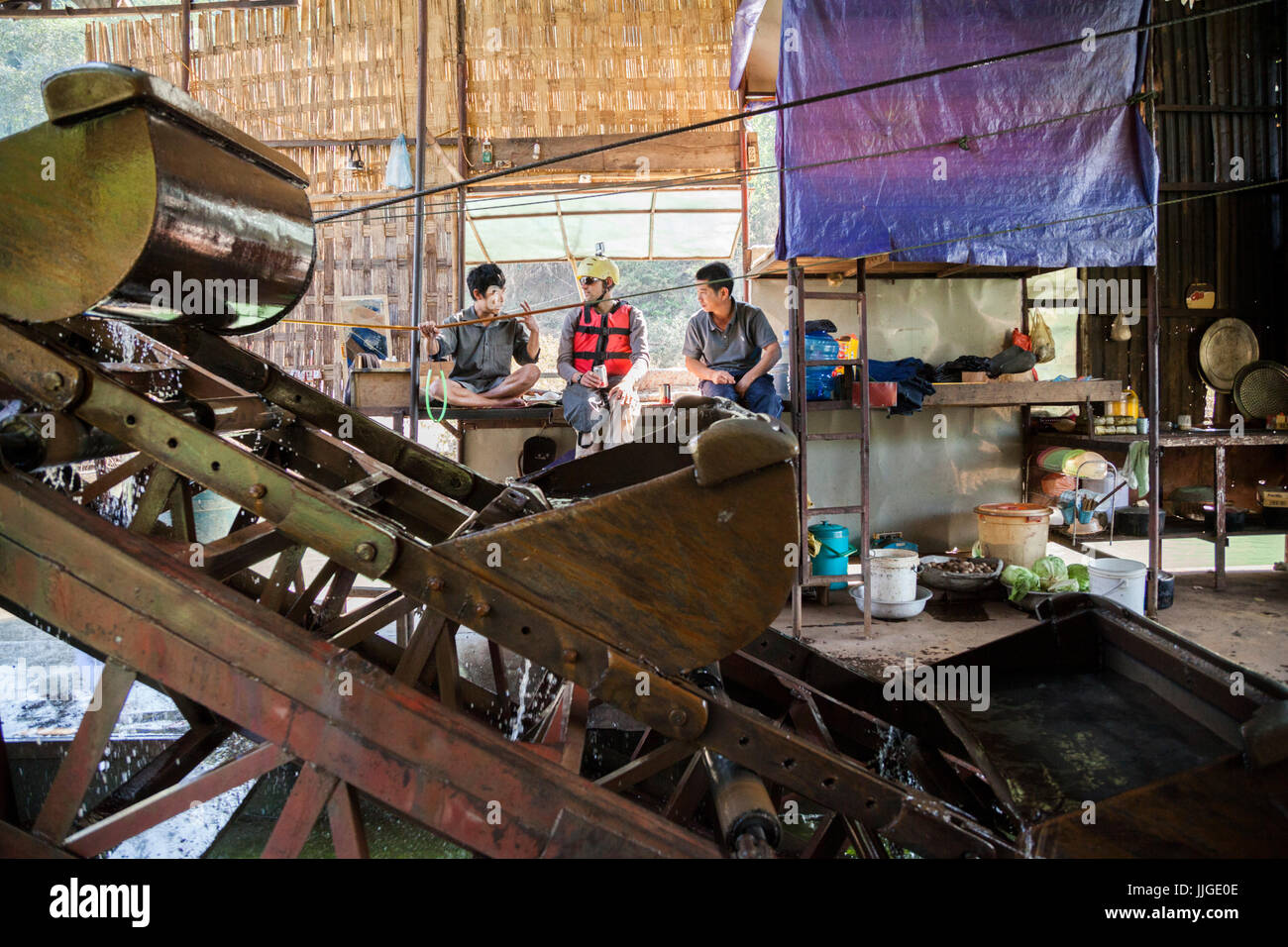 Robert Hahn (center) chats with Vietnamese workers, while drinking a Vietnamese beer, on a mechanical gold dredge floating on the Nam Ou River, Laos. Six men live on the vessel for months, rotating throughout the day and working in pairs to keep the machine in constant operation. Stock Photo