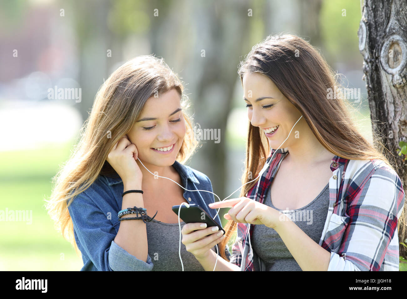 Two happy teen friends listening music sharing earphones and smartphone in a park Stock Photo