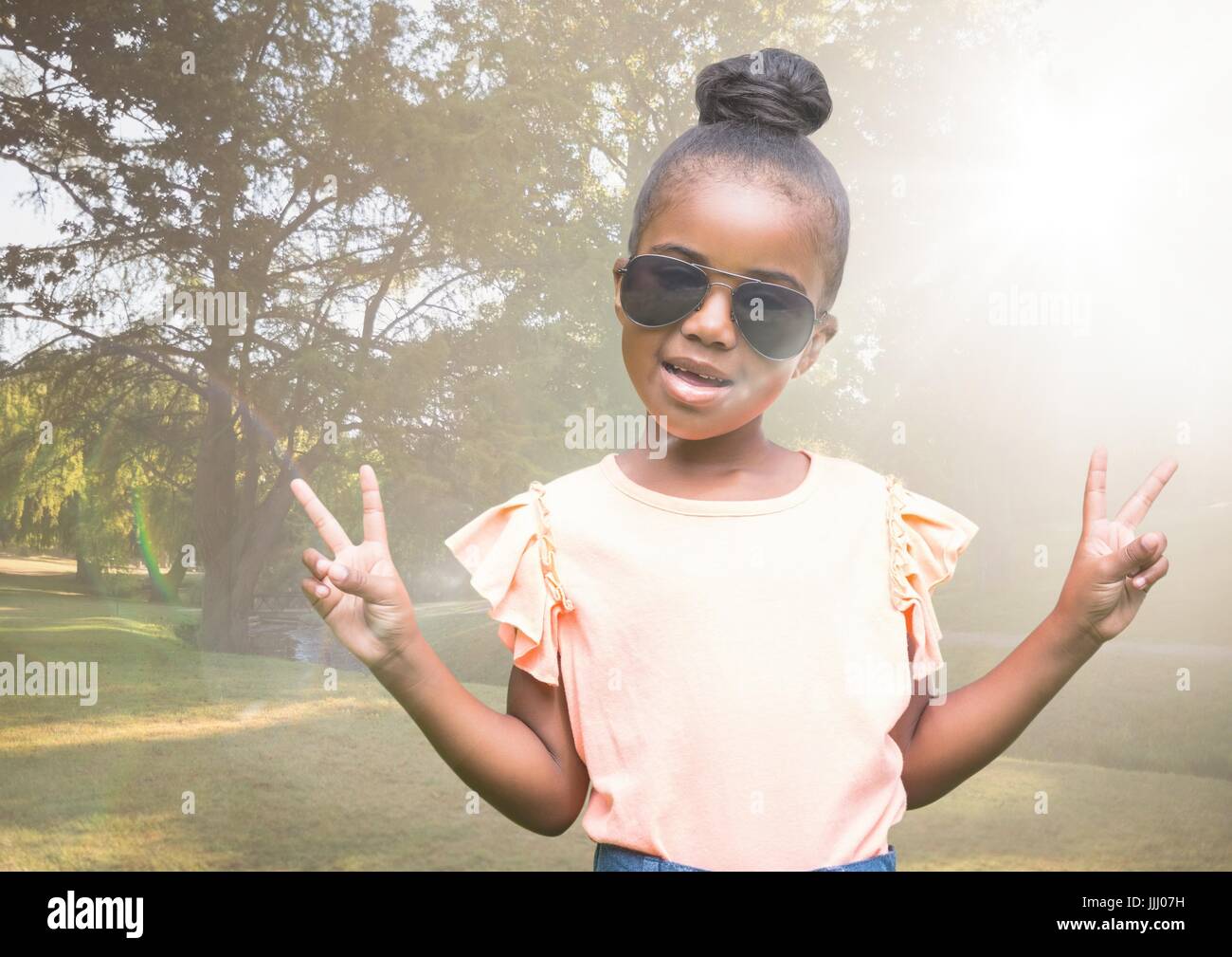 Girl in sunglasses making peace signs against blurry park with flare Stock Photo