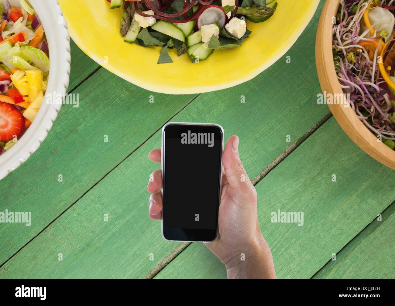 Person holding a phone against a green wooden table with food and copy space on mobile phone Stock Photo