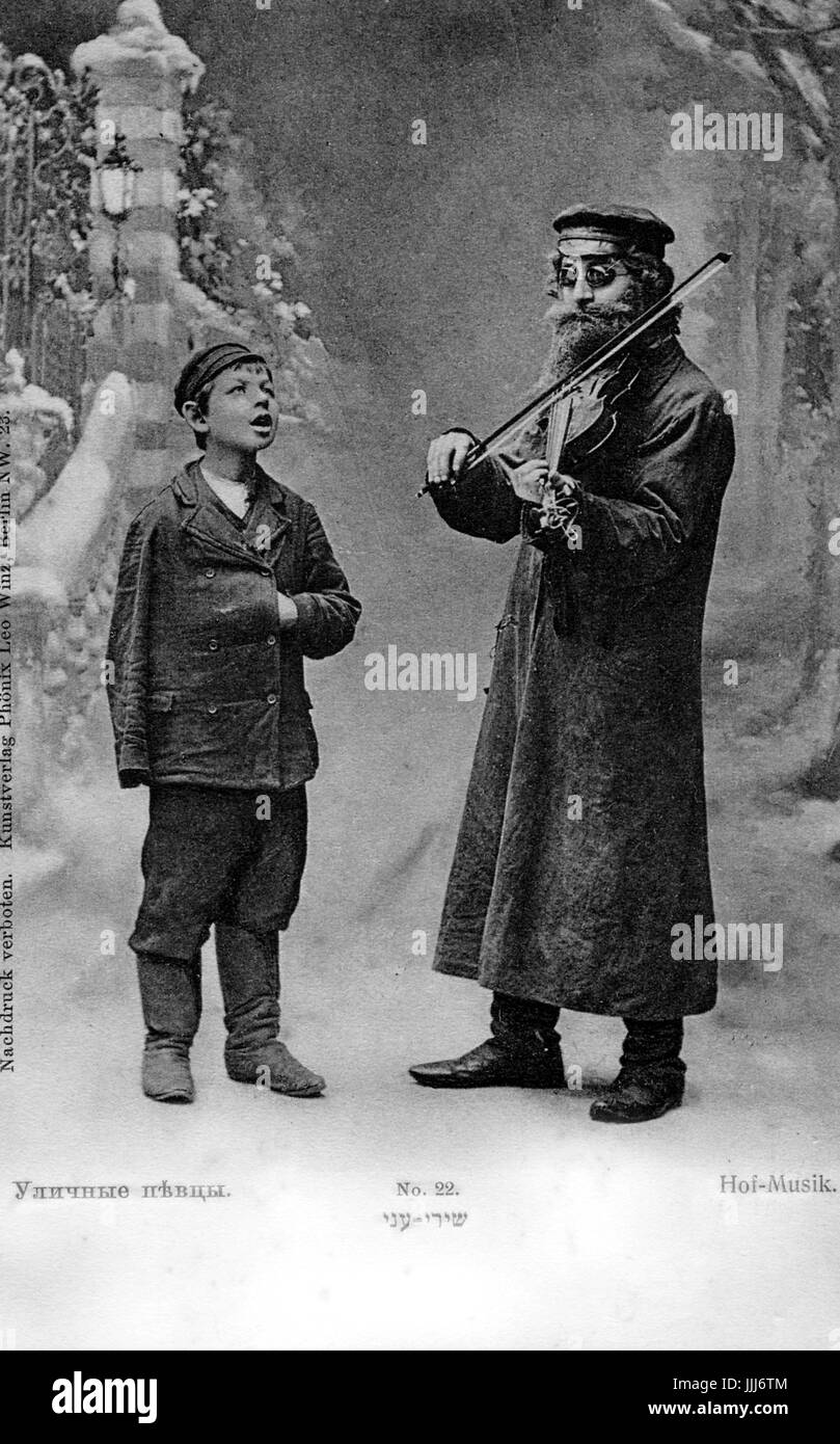 Jewish Musician with fiddle and boy singer. Shot against studio backdrop. Boy has one empty sleeve and hand inside his jacket.  Fiddle player wearing dark glasses, presumably blind.  Caption reads 'Hof-musik or in Yiddish 'Songs of the poor'.  Early 20th century. Stock Photo