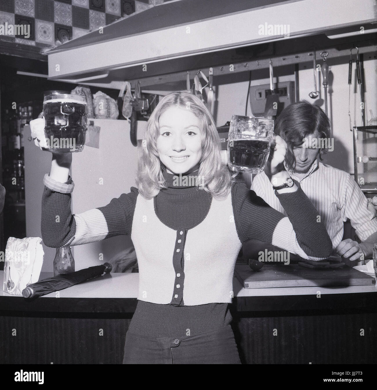 1970s, England, rag week celebrations, a young blonde female student wearing a tank-top holds up two pints of bitter. Stock Photo