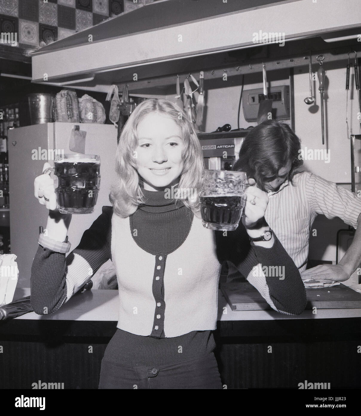 1970s, England, rag week celebrations, a young blonde female student wearing a tank-top holds up two pints of bitter. Stock Photo