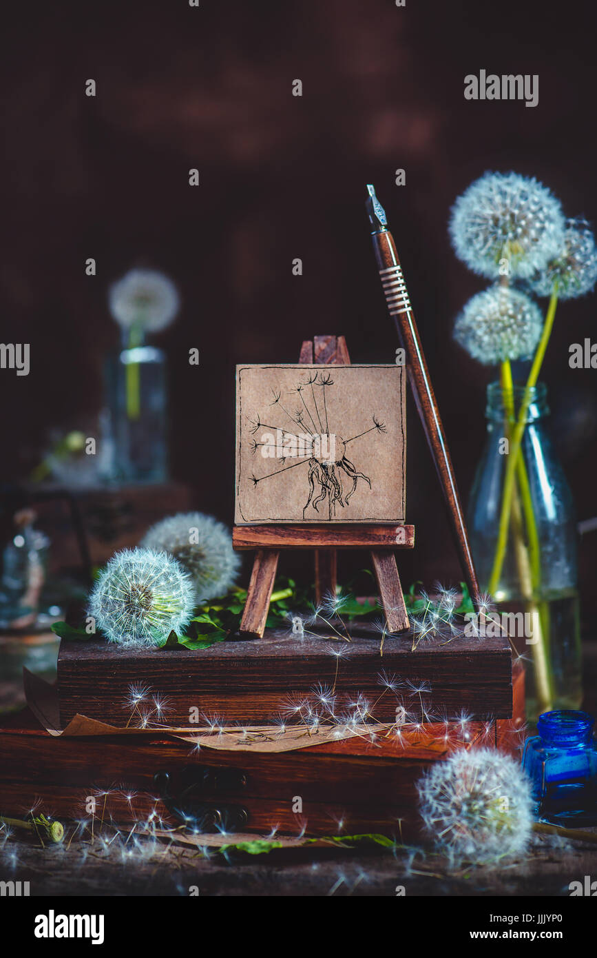 Summer still life with blowballs, a little dandelion painting on an easel, ink pen and, stack of wooden boxes on a dark wooden background Stock Photo