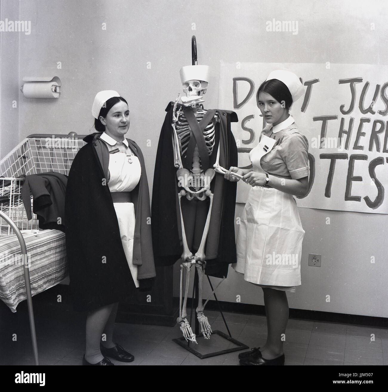 'Is it still alive?' 1960s, England, on a hospital ward, two female nurses with a life size model skeleton dressed with a nurse's cape and hat and a sandwich in its mouth. One of the nurse's pretends to check the pulse of the skeleton in a protest about surviving on low wages. Stock Photo