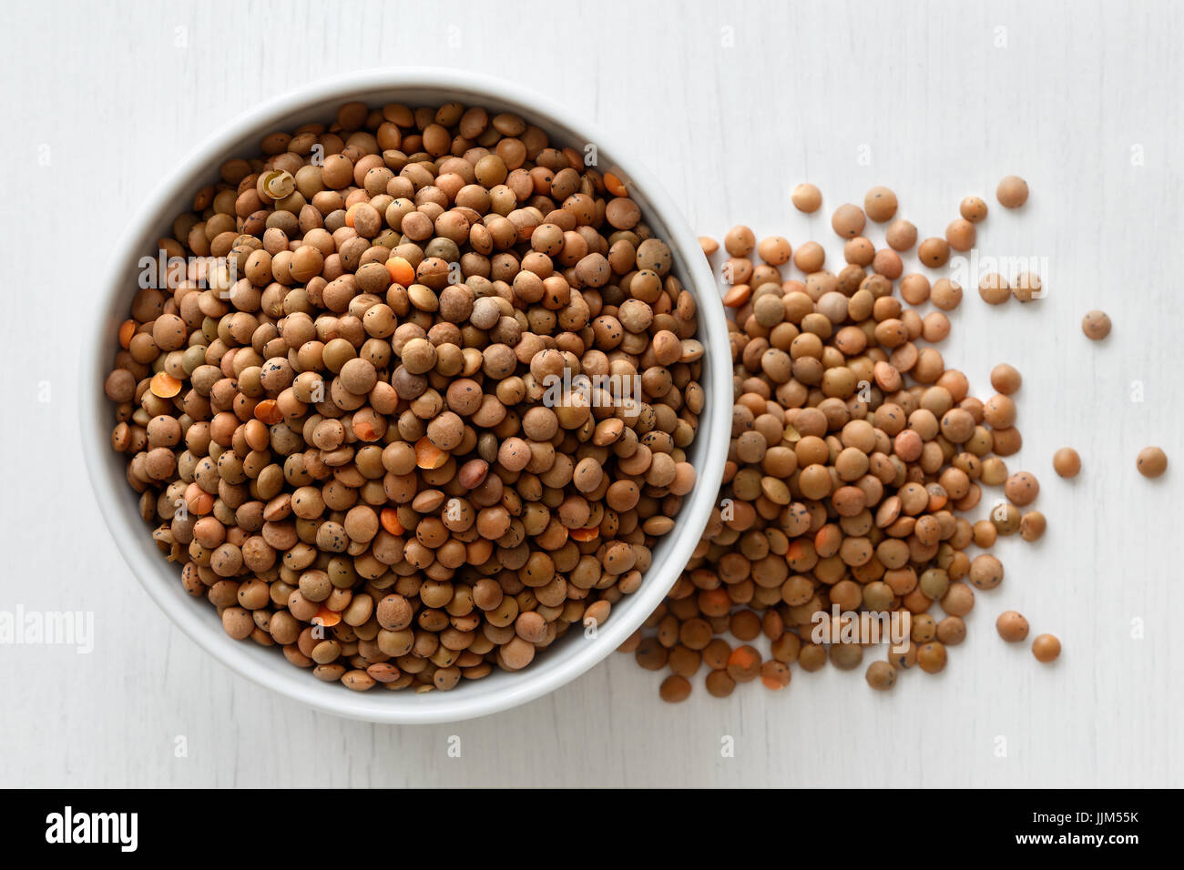 Dry unpeeled red lentils in white ceramic bowl isolated on painted white wood from above. Spilled lentils. Stock Photo