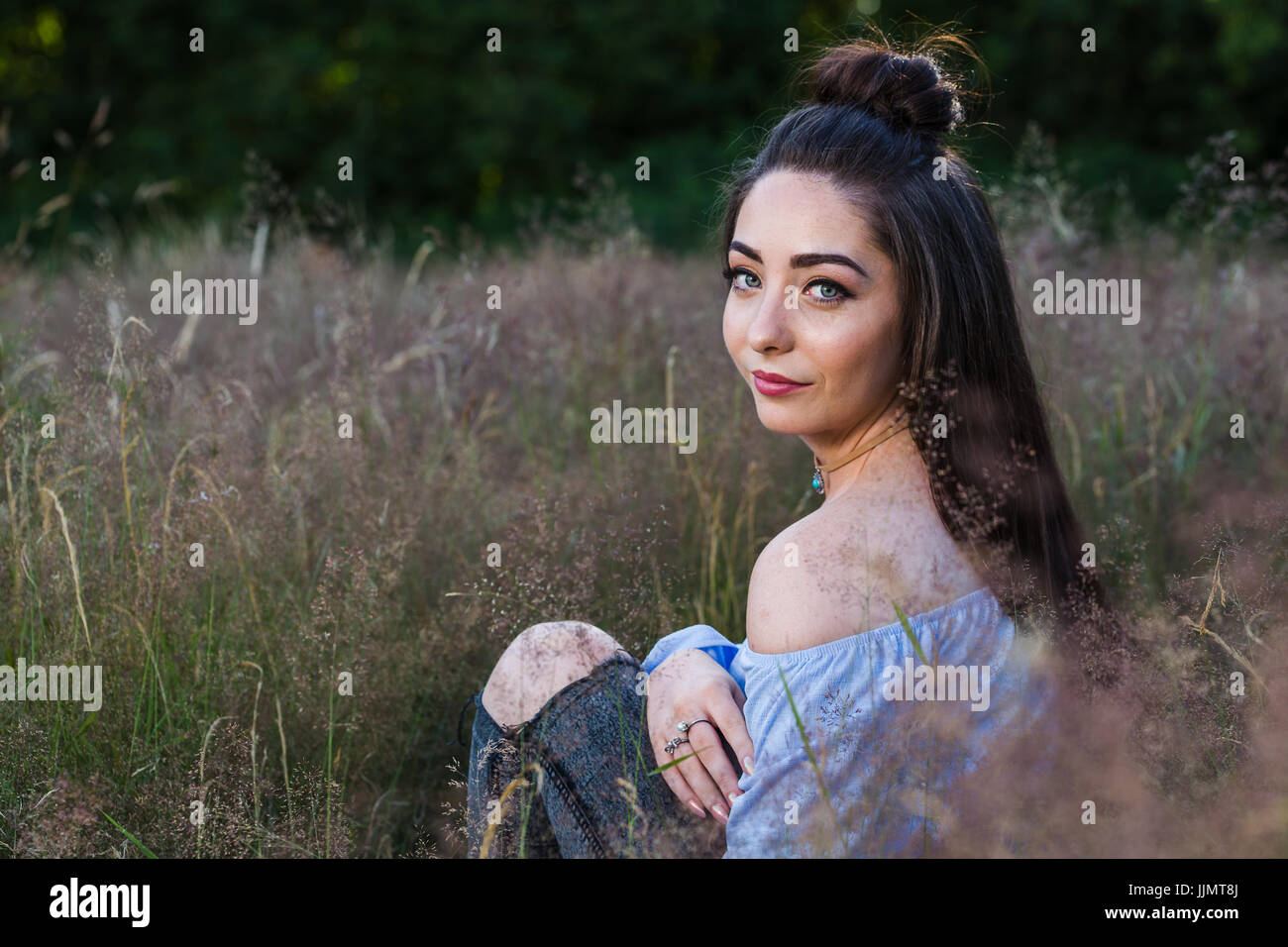 My friend looks over her shoulder whilst posing for a portrait in a grassy meadow near Liverpool during a summer evening. Stock Photo
