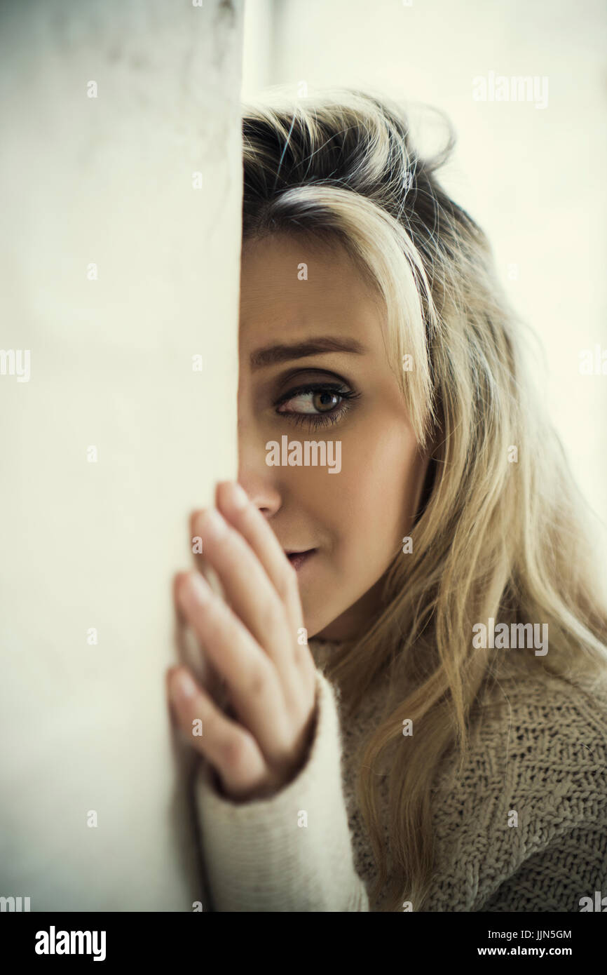 Scared young woman hiding behind wall looking away crying Stock Photo