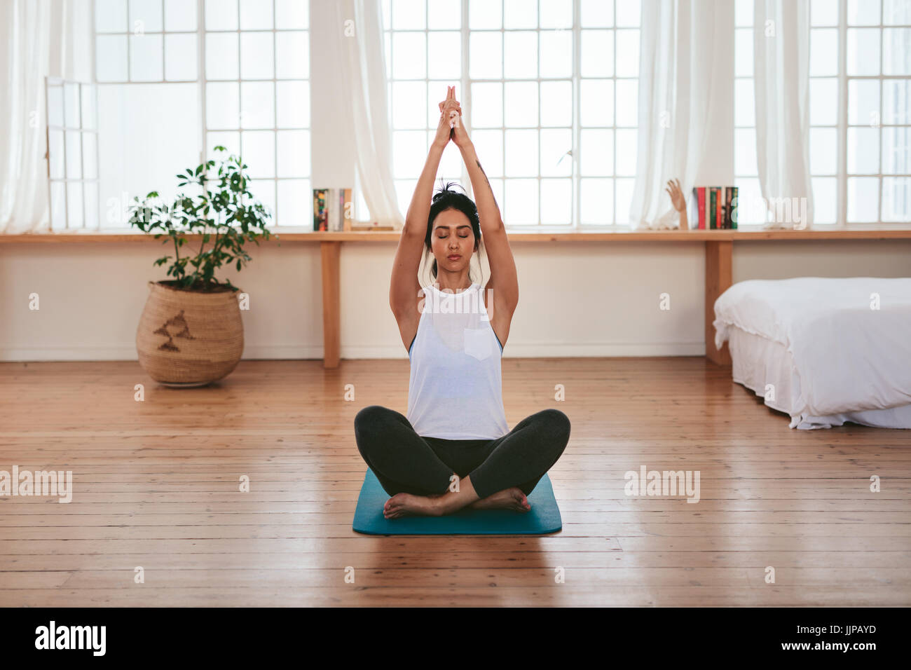 Full length shot of healthy and beautiful young woman sitting at home in yoga position. Fitness female sitting with hands joined above her head. Stock Photo