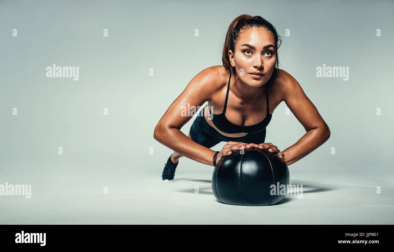 Horizontal shot of young fit woman doing push up on medicine ball. Fitness female exercising with a medicine ball on grey background. Stock Photo