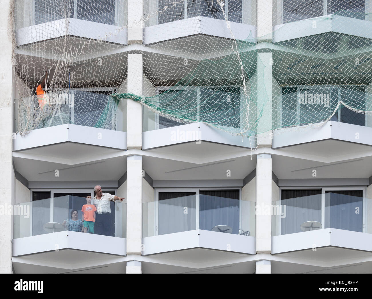 Workmen refurbishing upper floors of hotel in Spain as tourists sit on balconies on lower floors. Stock Photo
