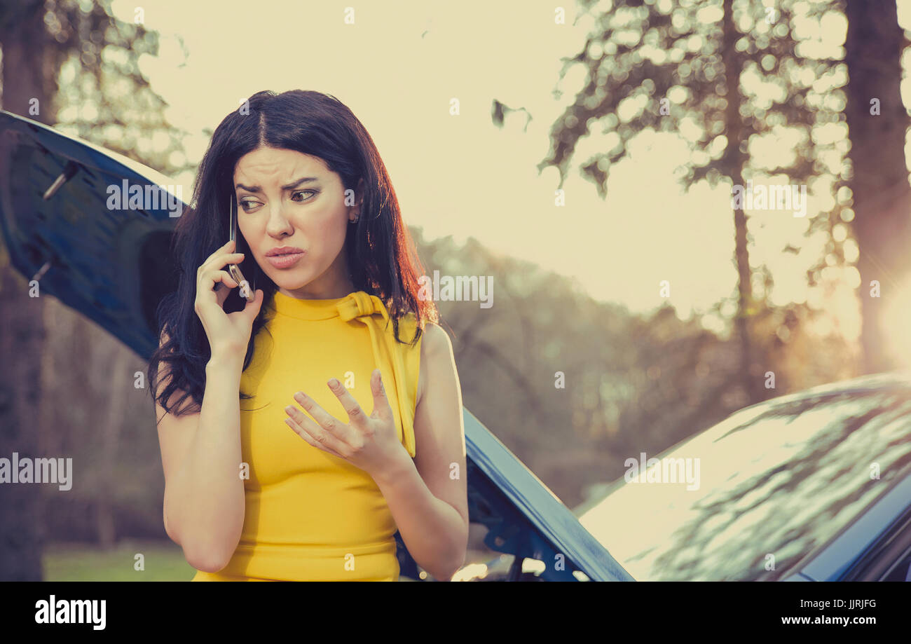 woman having trouble with her broken car, opening hood and calling for help on cell phone, isolated green trees and park background Stock Photo