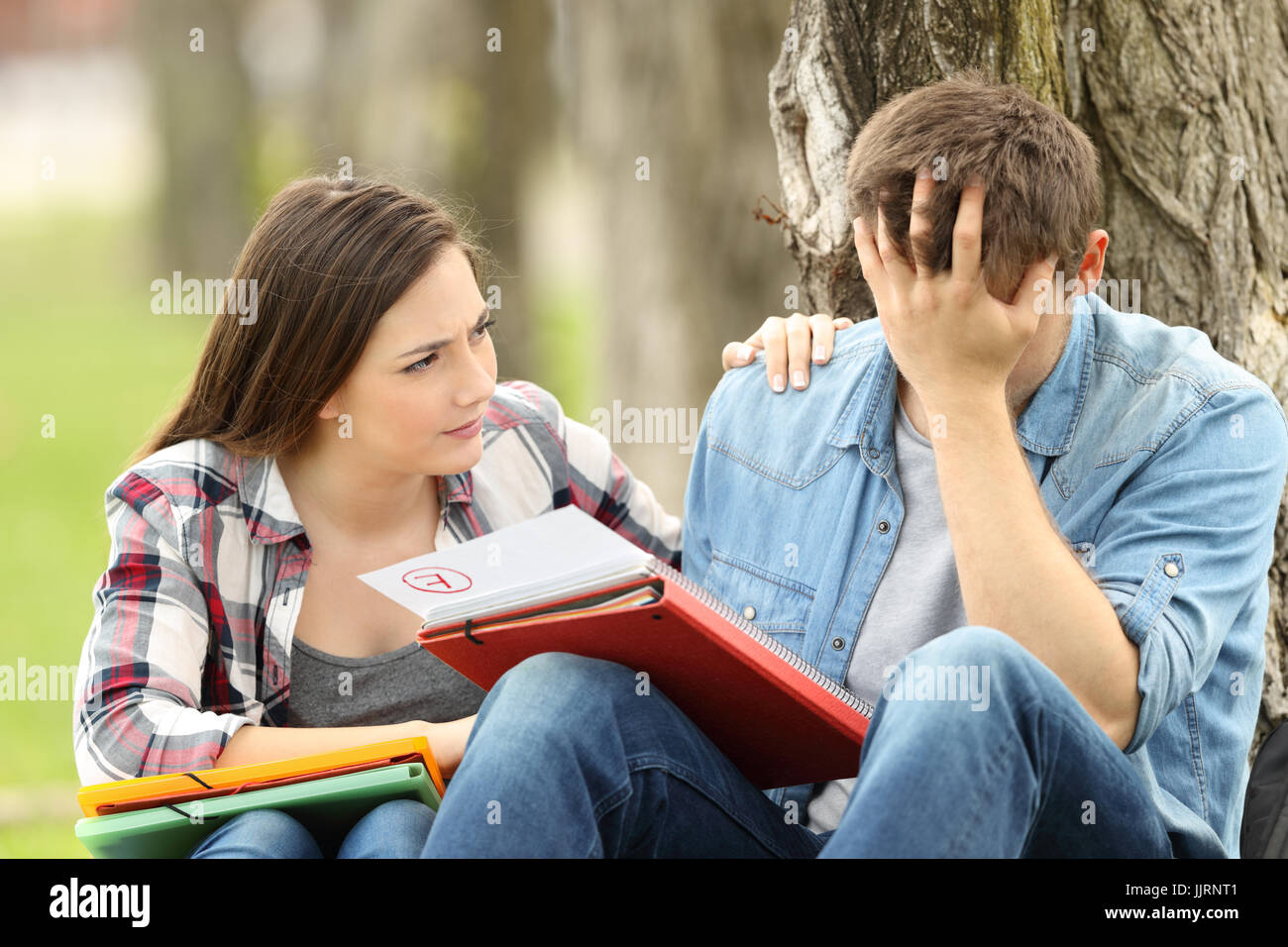 Friend comforting to a sad student with failed exam sitting on the grass in a park Stock Photo