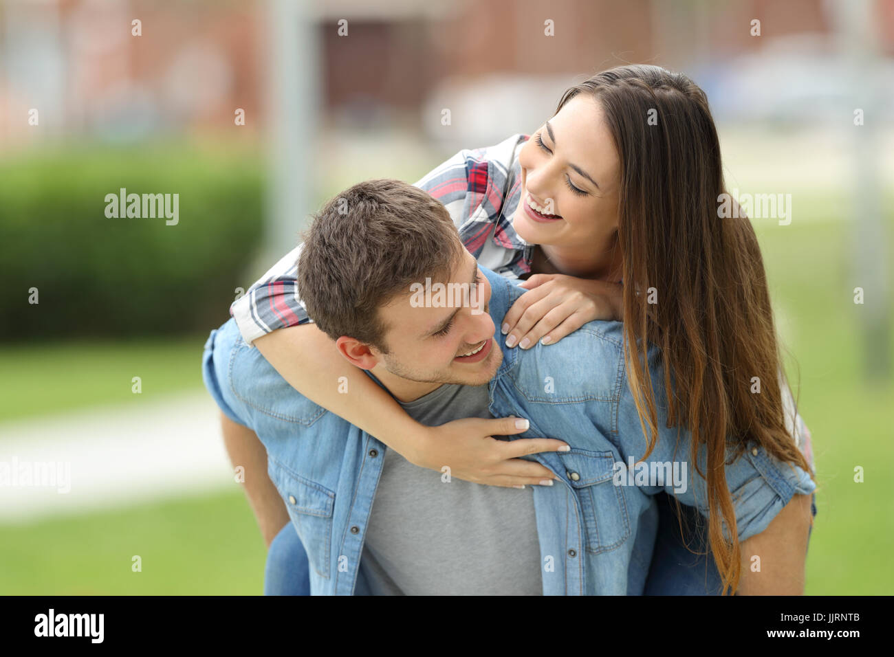 Front view of a happy couple of teens joking together piggyback outdoors in a park with a green background Stock Photo