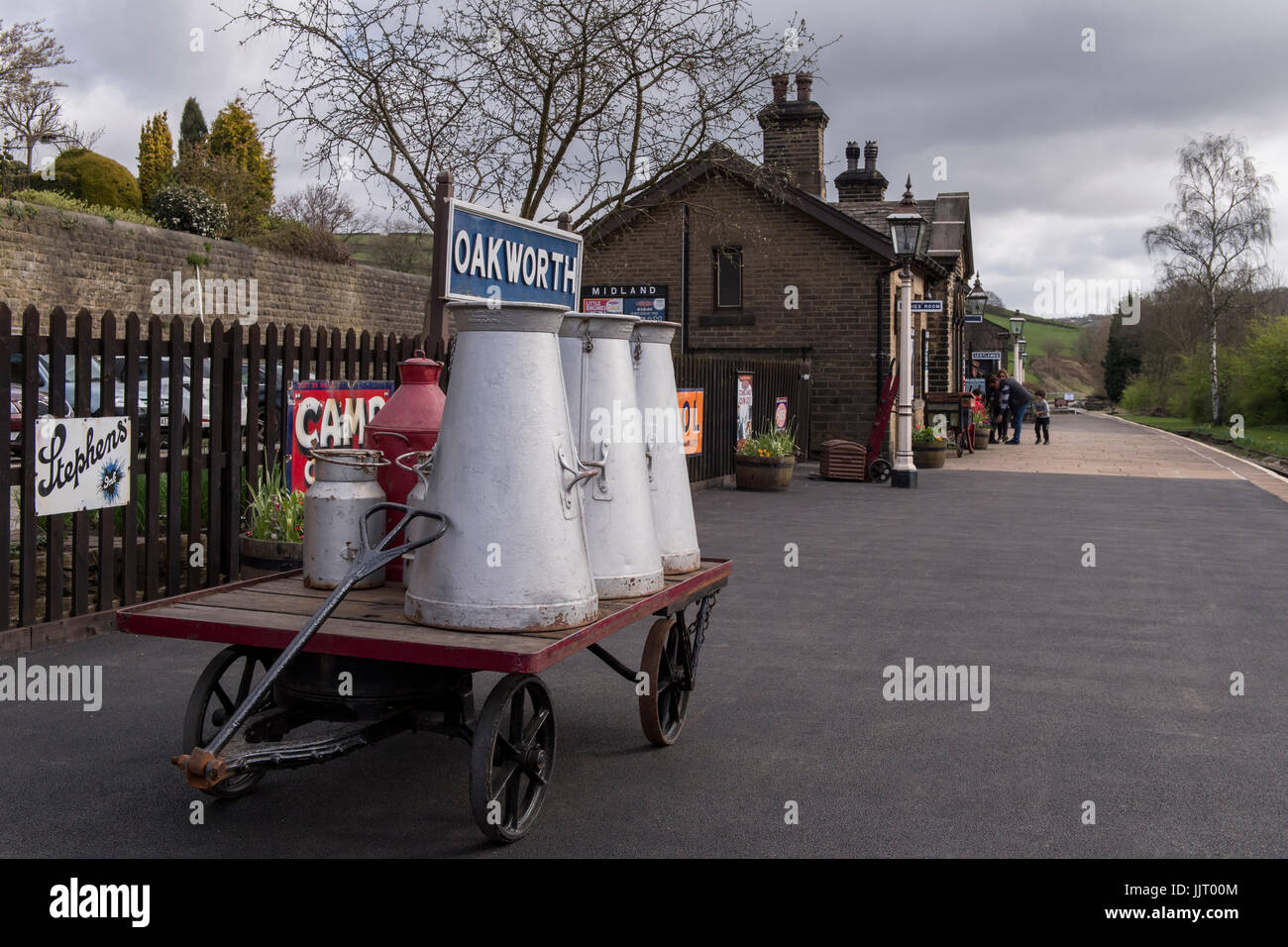 View of old adverts & milk churns on cart, on platform at quiet, quaint, historic Oakworth Station - Keighley and Worth Valley Railway, England, UK. Stock Photo